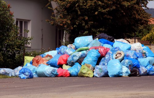 A large pile of trash bags is stacked along the edge of a residential street in St Johns Wood, London. The bags are made of plastic, predominantly in shades of blue, with some red, orange, green, and black, and appear to contain household or garden waste. They are positioned on the pavement and partially extend onto the road, with some bags leaning against each other for support. In the background, there is a white building with a window partially obscured by foliage, including a tree with dense, dark green leaves and reddish-brown branches. The scene suggests a rubbish removal service activity in the area, with Commercial Waste St Johns Wood likely responsible for managing the waste. The environment appears calm, with natural lighting indicating daytime, and the waste collection is temporarily assembled on the street, awaiting removal or disposal.