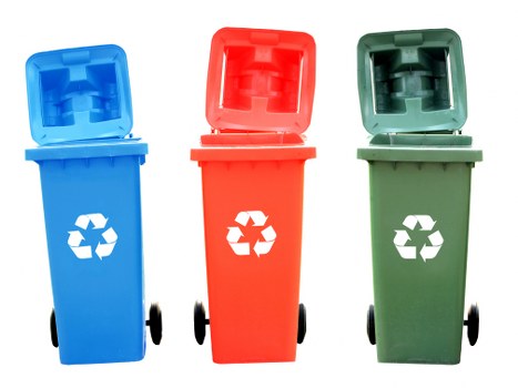 The image displays three individual wheeled rubbish bins made of durable plastic, arranged in a row on a plain white background. The first bin on the left is blue with a hinged lid open, revealing a textured interior surface, and features a white recycling symbol on the front. The middle bin is red, also with a raised lid, and similarly displays the white recycling emblem; it has a smooth matte finish and is positioned slightly behind the other two. The third bin on the right is green with its lid fully open, showing a similar textured interior, and carries the same recycling symbol in white. Each bin has black wheels at the base and is aligned evenly, suggesting they are ready for collection or disposal. The straightforward arrangement and neutral background focus attention on the different coloured waste bins, typical of those used for segregating rubbish and recyclable waste in urban environments, including areas around St Johns Wood, aligned with commercial waste management practices by companies like Commercial Waste St Johns Wood.