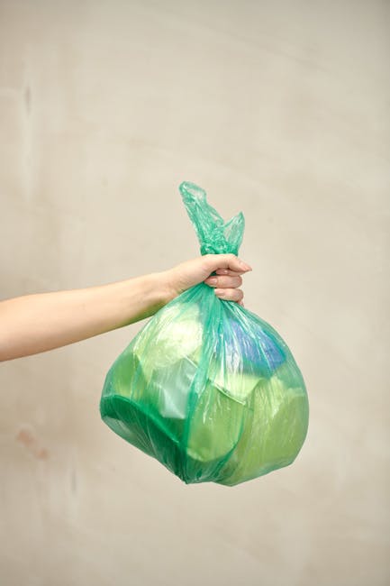 A person’s outstretched arm holding a green plastic rubbish bag with a knotted top, made of semi-transparent material revealing some contents inside. The bag appears to be filled with waste and is s