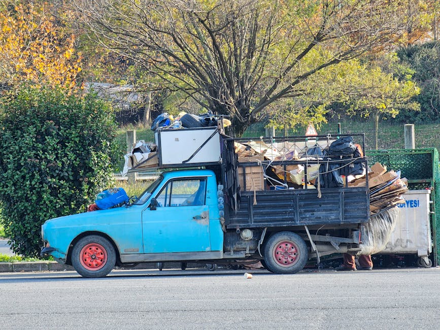 A waste collection vehicle with an open rear hopper, showing rusted metal surfaces and mechanical components, is parked on a narrow cobblestone street alongside old residential buildings with weathere