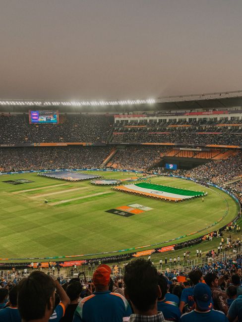 A large cricket stadium filled with spectators during a match, with the pitch and outfield visible in the centre. The field features a prominent cricket pitch at the middle, surrounded by a well-maint